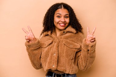 Genç Afrikalı Amerikalı kadın bej arka planda izole edilmiş zafer işareti gösteriyor ve gülümsüyor..