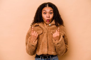 Genç Afrikalı Amerikalı kadın, bej arka planda izole edilmiş, ağzı açık, üst tarafı işaret ediyor..