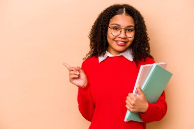 Genç Afrikalı Afrikalı Amerikalı kadın bej arka planda izole edilmiş gülümseyen ve işaret eden boş alanda bir şey gösteriyor..