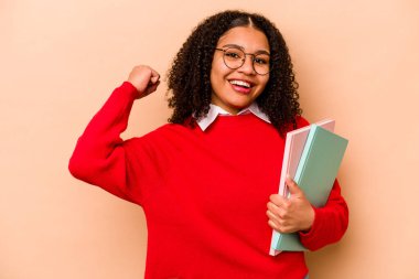 Genç Afrikalı Afrikalı Amerikalı kadın bej arka planda izole edilmiş zafer ve galibiyet kavramından sonra yumruğunu kaldırıyor..