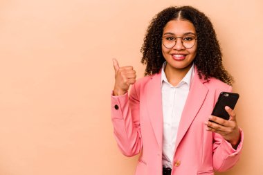 Elinde cep telefonu olan genç Afrikalı bir kadın bej arka planda izole edilmiş gülümsüyor ve başparmağını kaldırıyor.