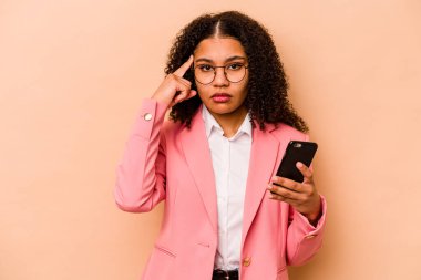 Elinde cep telefonu olan genç Afrikalı bir kadın bej arka planda izole edilmiş. Şakağı işaret ediyor, düşünüyor ve bir göreve odaklanıyor..