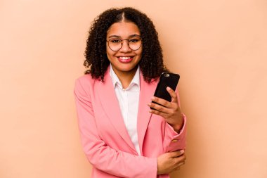 Elinde cep telefonu olan genç Afrikalı bir kadın bej arka planda izole edilmiş gülüyor ve eğleniyor..
