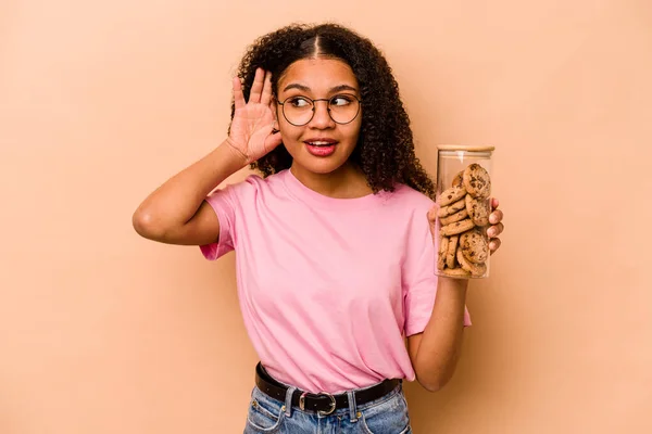 Young African american woman holding a cookies jar isolated on beige background trying to listening a gossip.