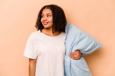 Genç Afrikalı Amerikalı bir kadın bej arka planda izole edilmiş bir yastık tutuyor. Gülümsüyor, neşeli ve keyifli görünüyor..