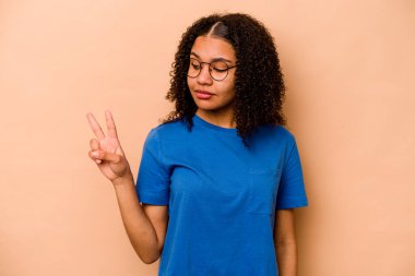 Genç Afrikalı Amerikalı kadın bej arka planda izole edilmiş neşeli ve kaygısız parmaklarıyla barış sembolü gösteriyor..