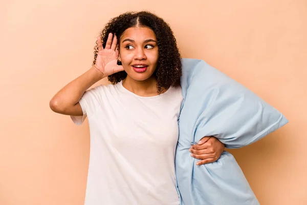 Young African American woman holding a pillow isolated on beige background trying to listening a gossip.