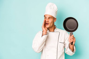 Kafkasyalı genç aşçı elinde uçan bir tavayla mavi arka planda izole edilmiş bir şekilde gizli bir sıcak fren haberi veriyor ve yana bakıyor.