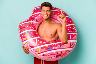 Mavi arka planda izole edilmiş şişirilebilir donut tutan genç beyaz bir adam bir numarayı parmağıyla gösteriyor..