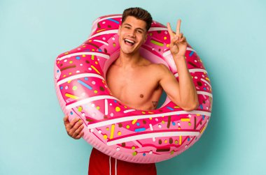 Mavi arka planda izole edilmiş şişme bir çörek tutan genç beyaz adam neşeli ve tasasız parmaklarıyla barış sembolü gösteriyor..