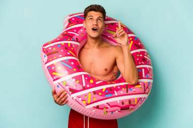 Genç beyaz adam mavi arka planda izole edilmiş şişirilebilir bir çörek tutuyor ve ağzı açık bir şekilde yukarı bakıyor..