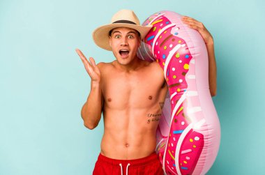 Mavi arka planda izole edilmiş şişme bir çörek tutan genç beyaz adam şaşırdı ve şok oldu..