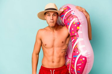 Mavi arka planda izole edilmiş şişme bir çörek tutan genç beyaz adam omuzlarını silker ve gözleri açık kalır..