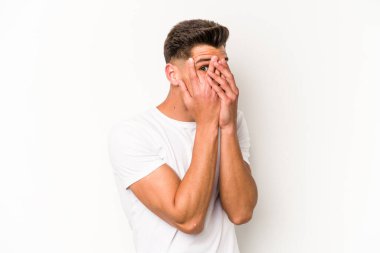 Beyaz arka planda izole edilmiş genç beyaz bir adam parmaklarının arasından korkmuş ve gergin bir şekilde göz kırpıyor..