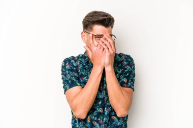 Beyaz arka planda izole edilmiş genç beyaz bir adam kameraya parmaklarıyla göz kırpıyor, yüzünü kapatmaktan utanıyor..