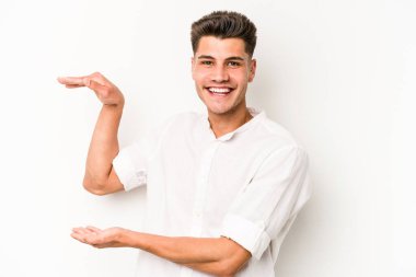 Beyaz arka planda izole edilmiş genç beyaz bir adam işaret parmaklarıyla küçük bir şey tutuyor, gülümsüyor ve kendinden emin..