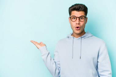 Mavi arka planda izole edilmiş genç beyaz adam, avuç içi fotokopi alanını elinde tutmaktan etkilenmiş..