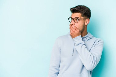 Kafkasyalı genç adam mavi arka planda izole edilmiş. Düşünceli bir şekilde ağzıyla ağzını kapatan bir fotokopi odasına bakıyor..