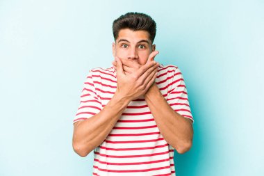 Mavi arka planda izole edilmiş genç beyaz adam elleriyle ağzını kapatırken şok oldu..