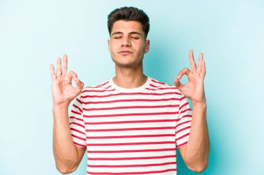 Mavi arka planda izole edilmiş genç beyaz adam yorucu bir günün ardından rahatlıyor. Yoga yapıyor..