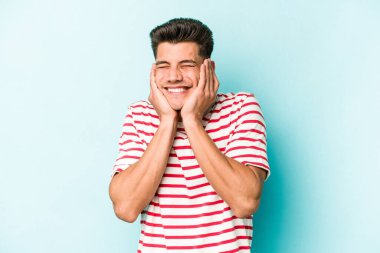 Mavi arka planda izole edilmiş genç beyaz adam kulaklarını elleriyle kapatıyor..