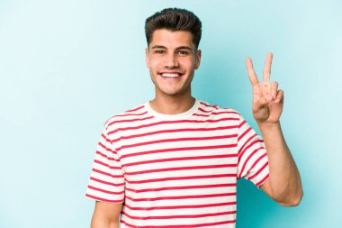 Kafkasyalı genç adam mavi arka planda izole edilmiş neşeli ve kaygısız parmaklarıyla barış sembolü gösteriyor..