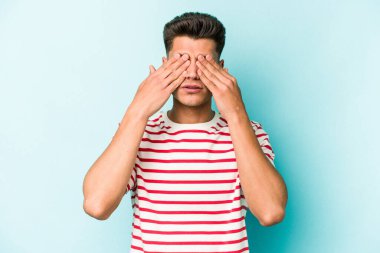 Mavi arka planda izole edilmiş genç beyaz adam elleriyle gözlerini kapatmaktan korkuyor..