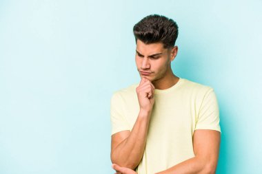 Mavi arka planda izole edilmiş genç beyaz adam şüpheli ve şüpheci bir ifadeyle yana bakıyor..