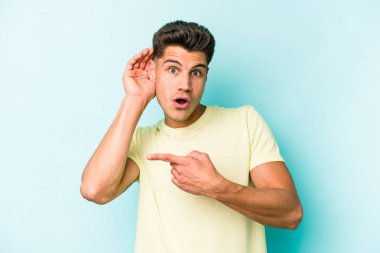 Mavi arka planda izole edilmiş genç beyaz adam bir dedikoduyu dinlemeye çalışıyor..