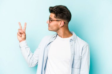 Kafkasyalı genç adam mavi arka planda izole edilmiş neşeli ve kaygısız parmaklarıyla barış sembolü gösteriyor..