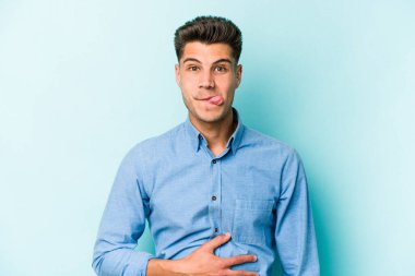 Mavi arka planda izole edilmiş genç beyaz adam karnına dokunuyor, nazikçe gülümsüyor, yemek yiyor ve tatmin oluyor..