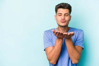 Kafkasyalı genç adam mavi arka planda izole edilmiş dudaklar kıvrılıyor ve hava öpücüğü göndermek için avuç içleri tutuyor..
