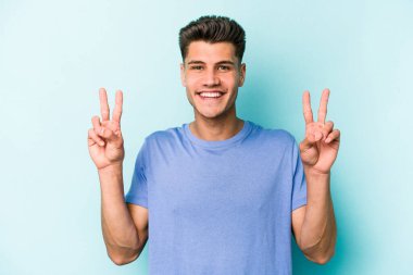 Mavi arka planda izole edilmiş genç beyaz adam zafer işareti gösteriyor ve gülümsüyor..