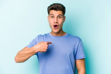 Mavi arka planda izole edilmiş genç beyaz adam parmağını göstererek şaşırdı, geniş bir gülümsemeyle..