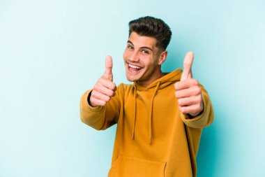 Kafkasyalı genç adam mavi arka planda izole edilmiş. Baş parmağını kaldırıyor, gülümsüyor ve kendinden emin..