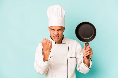 Kafkasyalı genç aşçı elinde uçan tavayla mavi arka planda izole edilmiş. Kameraya yumruk, agresif yüz ifadesi gösteriyor..