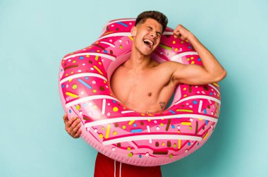 Mavi arka planda izole edilmiş şişirilebilir donut tutan genç beyaz adam zafer kazandıktan sonra yumruğunu kaldırıyor..