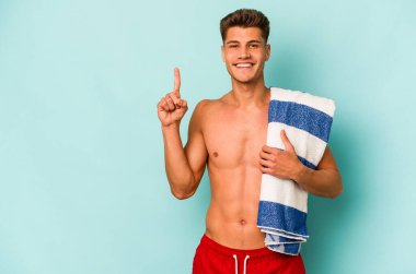 Elinde plaj havlusu tutan beyaz bir adam mavi arka planda bir numarayı parmağıyla gösteriyor..
