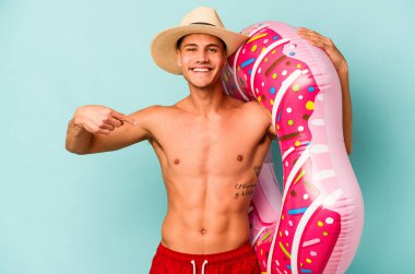 Mavi arka planda izole edilmiş şişme donut tutan genç beyaz adam gömlek fotokopi alanını işaret ediyor, gururlu ve kendinden emin.