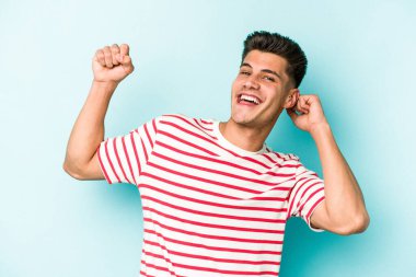 Mavi arka planda izole genç beyaz adam özel bir günü kutluyor, atlıyor ve enerjiyle kollarını kaldırıyor..