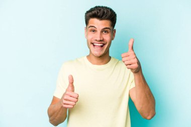 Kafkasyalı genç adam mavi arka planda izole edilmiş. Baş parmağını kaldırıyor, gülümsüyor ve kendinden emin..