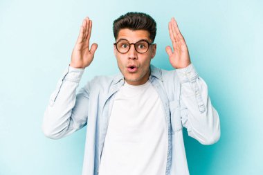 Mavi arka planda izole edilmiş genç beyaz bir adam zaferi ya da başarıyı kutluyor, şaşırıyor ve şok oluyor..