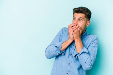 Kafkasyalı genç adam mavi arka planda izole edilmiş. Düşünceli bir şekilde ağzıyla ağzını kapatan bir fotokopi odasına bakıyor..