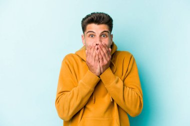 Mavi arka planda izole edilmiş genç beyaz adam şok olmuş, ağzı ellerle kaplanmış, yeni bir şey keşfetmeye hevesli..