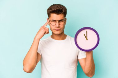 Young caucasian man holding a clock isolated on blue background pointing temple with finger, thinking, focused on a task.