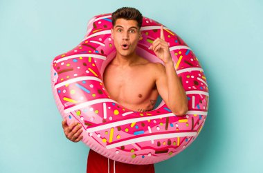 Mavi arka planda izole edilmiş şişirilebilir donut tutan genç beyaz bir adam harika bir fikre, yaratıcılık kavramına sahip..