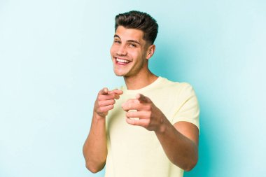 Mavi arka planda izole edilmiş genç beyaz adam parmaklarıyla ön tarafı işaret ediyor..