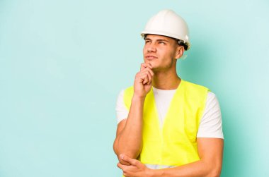 Kafkasyalı genç işçi mavi arka planda izole edilmiş şüpheli ve şüpheci bir ifadeyle yana bakıyor..