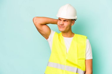 Kafkasyalı genç işçi mavi arka planda izole edilmiş kafasının arkasına dokunuyor, düşünüyor ve bir seçim yapıyor..