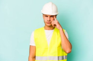 Kafkasyalı genç işçi, mavi arka planda izole edilmiş. Şakağı işaret ediyor, düşünüyor, bir göreve odaklanıyor..